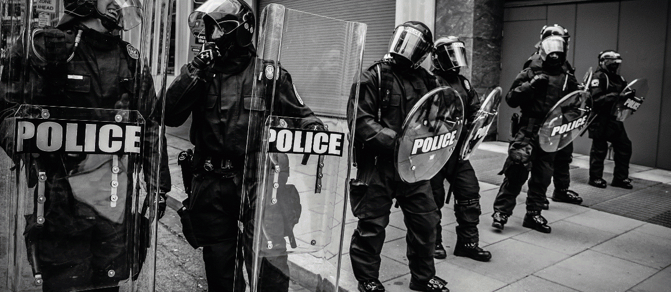 a black and white image of seven police officers with riot guards all stood in a line
