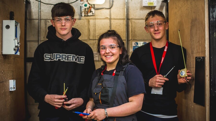 Three Barnsley College students with protective glasses on and tape measures and drawing materials in their hands, inside a workshop.