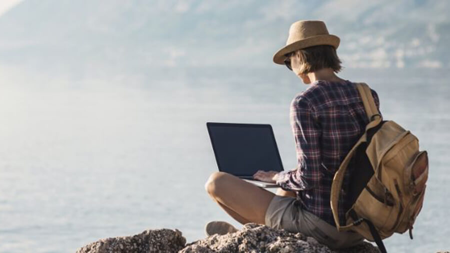 Woman sat on a rock at the coast working on her laptop wearing a sunhat and glasses