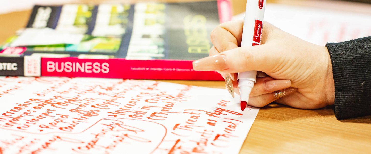 A hand making notes with a thick marker pen on a large piece of paper. There is a textbook entitled 'business' in the background.
