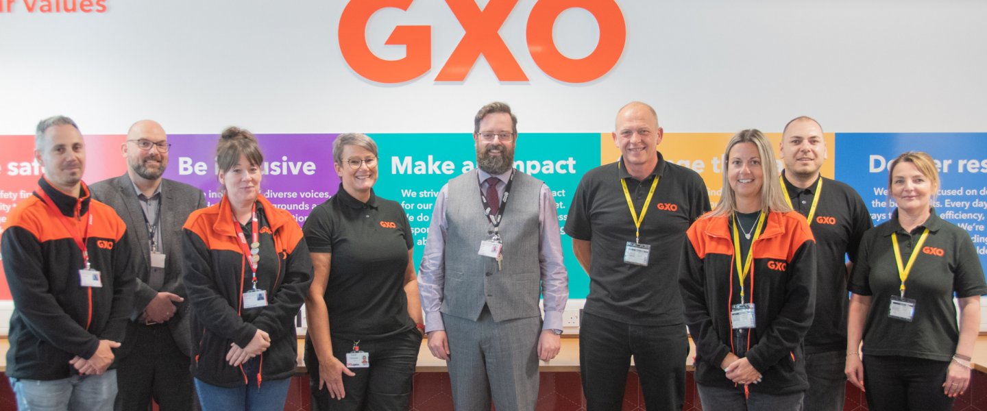 A group of men and women stand smiling in front of an orange sign that reads 'GXO'