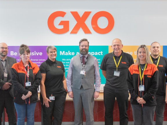 A group of men and women stand smiling in front of an orange sign that reads 'GXO'