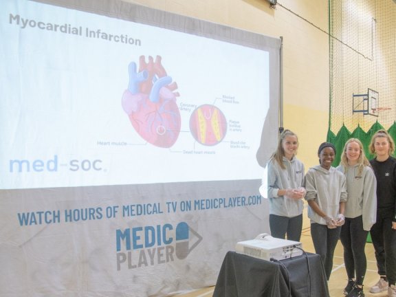 A group of students stand in front of a large screen with a diagram of a heart and the words 'myocardial infarction'