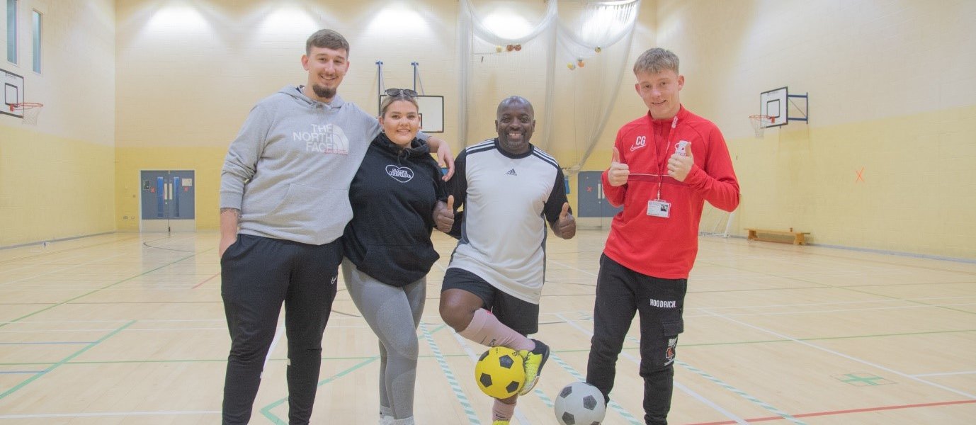 Three students dressed in sports wear pose with a footballer, who is balancing a football on his foot