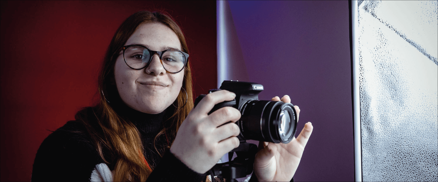 A girl with red hair holds a DSLR camera and smiles