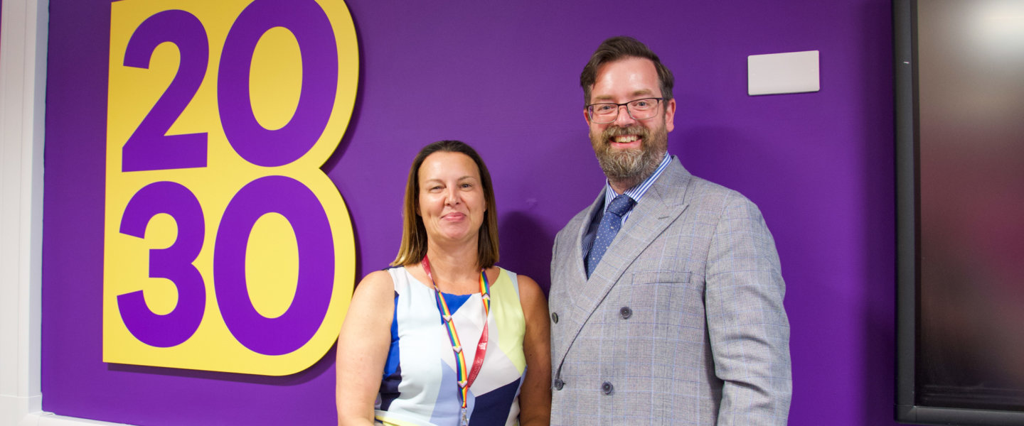 A woman with brown hair in her 40s stands next to a man in his 50s with glasses and a beard in front of a purple wall with yellow writing which says 'Barnsley 2030 - the place of possibilities'.