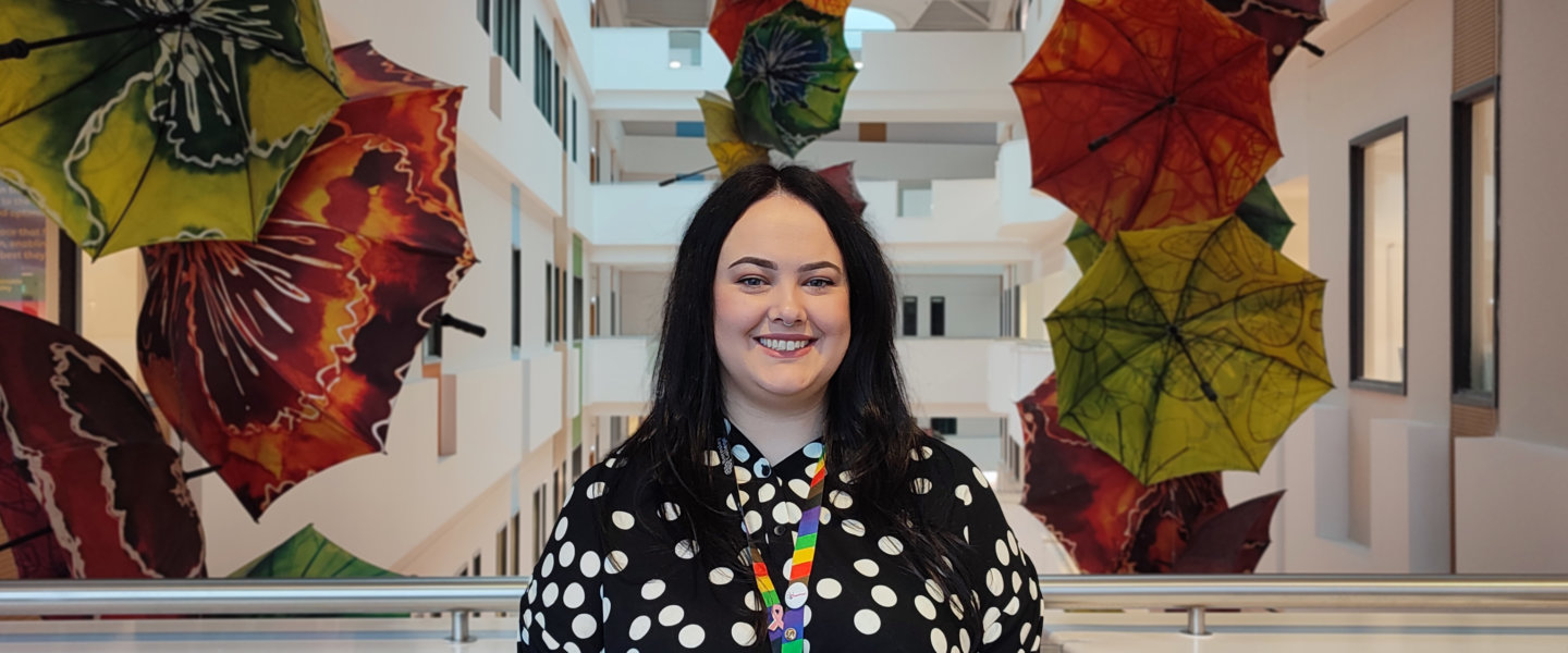 Sasha Beswick, Sustainability Officer at Barnsley College, stands on an indoor balcony with a backdrop of coloured umbrellas hung from the ceiling