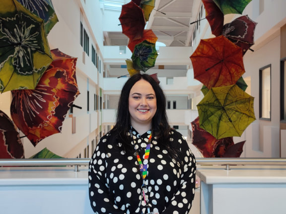 Sasha Beswick, Sustainability Officer at Barnsley College, stands on an indoor balcony with a backdrop of coloured umbrellas hung from the ceiling