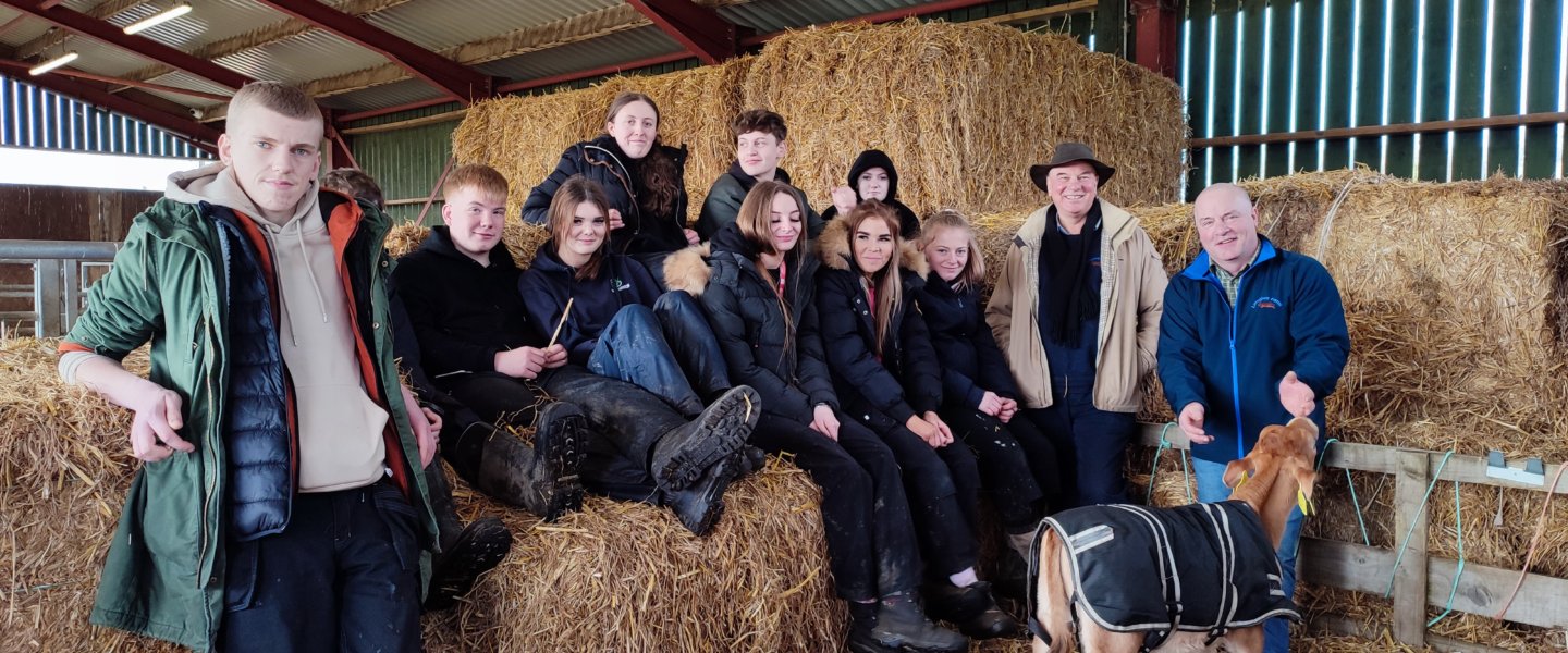 A group of students sit on haybales in a farm building, alongside two farmers, one of which is petting a young cow. Longley Farm's Jimmy Dickinson and Paul Robson with Agriculture students at Wigfield Farm.