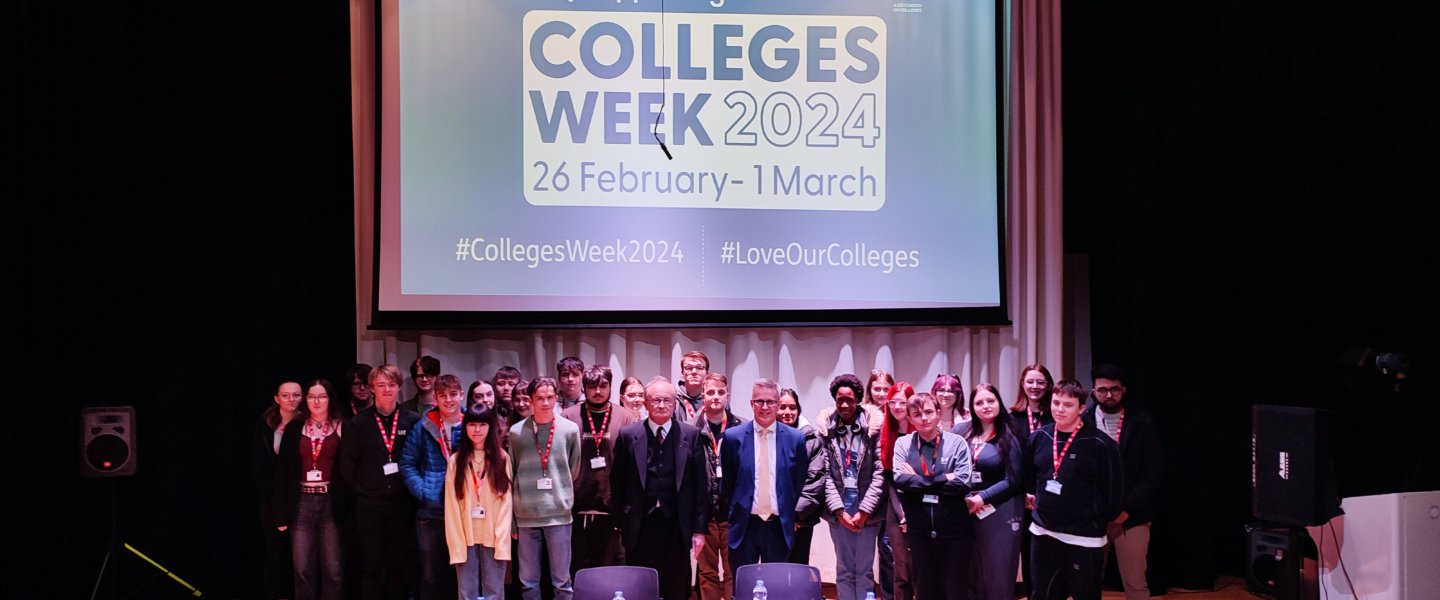 A group of students stand in a lecture theatre with two local councillors. In front of them is a table, at which the councillors were previously sitting. Behind the large group is a projection of Colleges Week 2024 graphics.