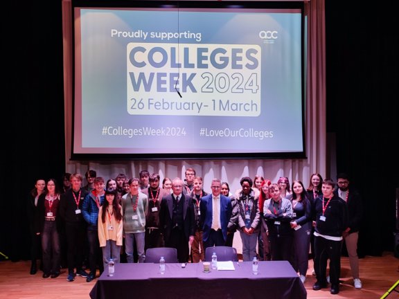 A group of students stand in a lecture theatre with two local councillors. In front of them is a table, at which the councillors were previously sitting. Behind the large group is a projection of Colleges Week 2024 graphics.