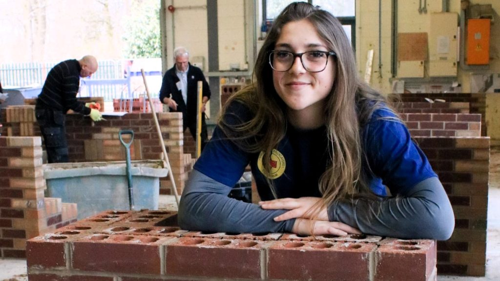 Female bricklaying apprentice from Barnsley College, Morgan Simmons, leans on a wall she has built at the Guild of Bricklayers competition. Behind her in the workshop are two other competitors also working on brickwork.