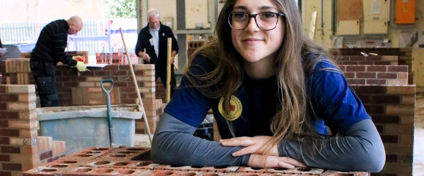 Female bricklaying apprentice from Barnsley College, Morgan Simmons, leans on a wall she has built at the Guild of Bricklayers competition. Behind her in the workshop are two other competitors also working on brickwork.