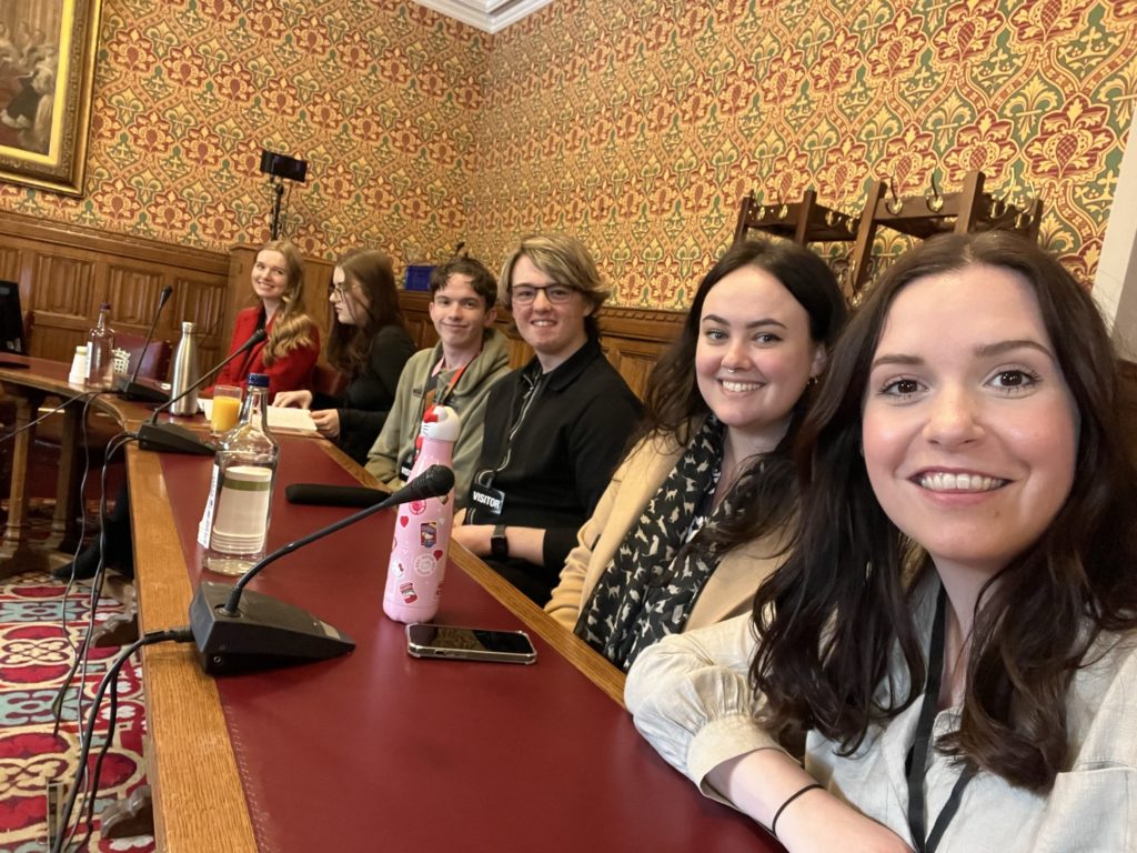 A group of students and staff sit at a long table in a room in the Houses of Parliament. They have microphones on the table in front of them alongside bottles of water. The person in the foreground is taking the selfie with the rest of the group alongside her.